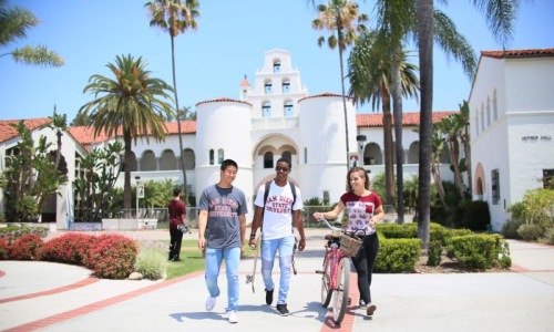 a group of people walking on a road with palm trees and a white building in the background with San Diego State University in the background