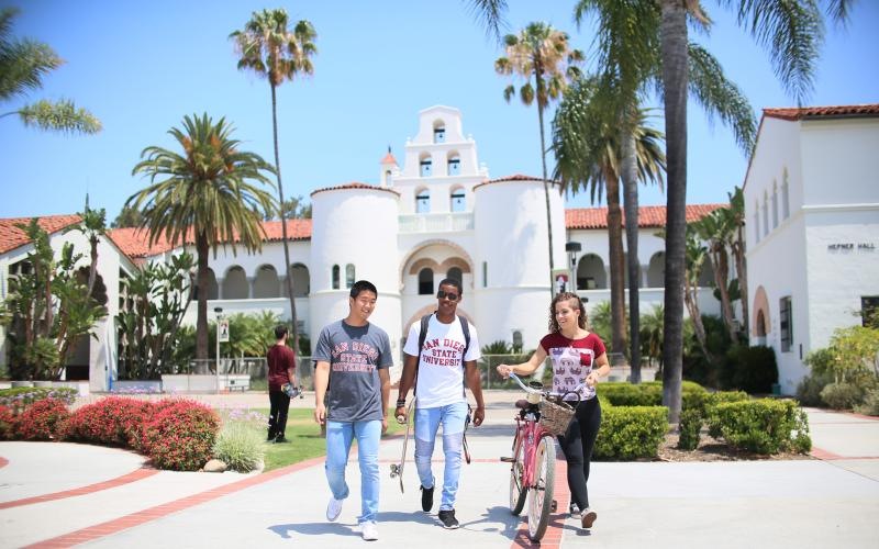 a group of people walking on a road with palm trees and a white building in the background with San Diego State University in the background